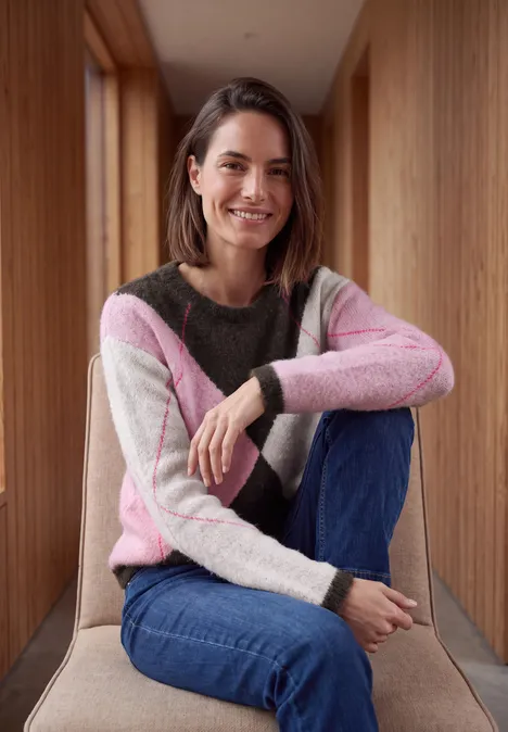 Pullover mit Rundhals und Argyle-Muster truffle brown Eine Frau sitzt in einem Holzflur, trägt einen bunten Pullover und Jeans, lächelt in die Kamera.