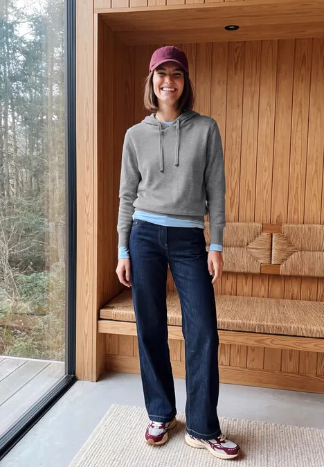 Personne en sweat gris, chemise bleue, jean foncé, baskets et casquette rouge dans un intérieur en bois