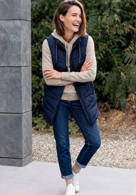 Lachende Frau in lässigem Outfit mit dunkelblauer Weste, Jeans und Sneakers lehnt an einer Säule im Garten.
