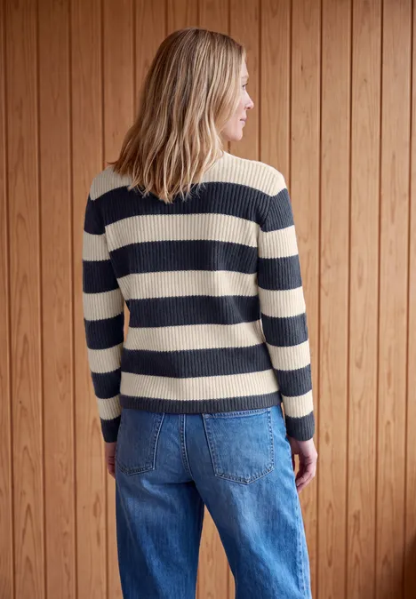 Femme de dos portant un pull rayé bleu et beige et un jean, devant un mur en bois.