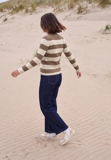 Une jeune femme en pull rayé et jean foncé se promène sur les dunes de sable près de la plage.