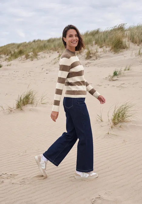 Femme souriante marchant sur la plage en pull rayé, jean large et baskets blanches.
