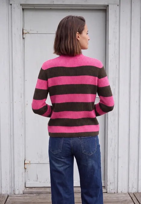 Une femme de dos porte un pull rayé rose et marron avec un jean bleu, devant une porte blanche.