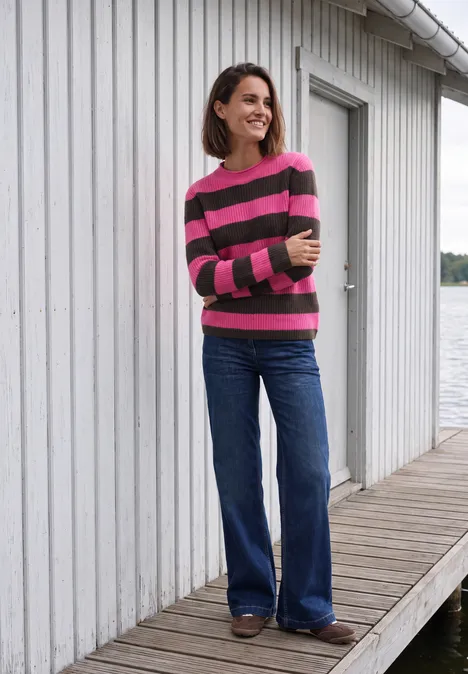 Une femme en pull rayé et jean large se tient sur une jetée devant une maison blanche avec une fenêtre.