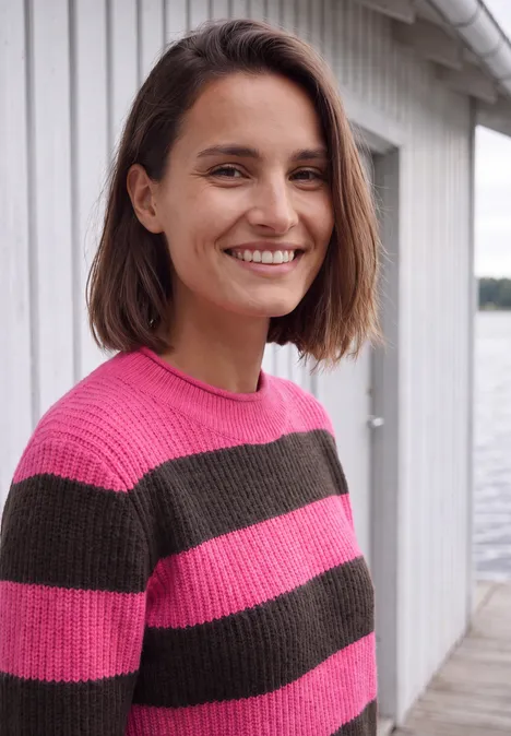 Une femme aux cheveux courts bruns sourit, vêtue d’un pull rayé rose et gris devant un fond clair.