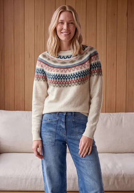 Pullover mit Rundhals und Jacquardmuster oat milk beige melange Lächelnde Frau trägt Fair-Isle-Pullover und Jeans, steht vor Holzvertäfelung und Sofa.