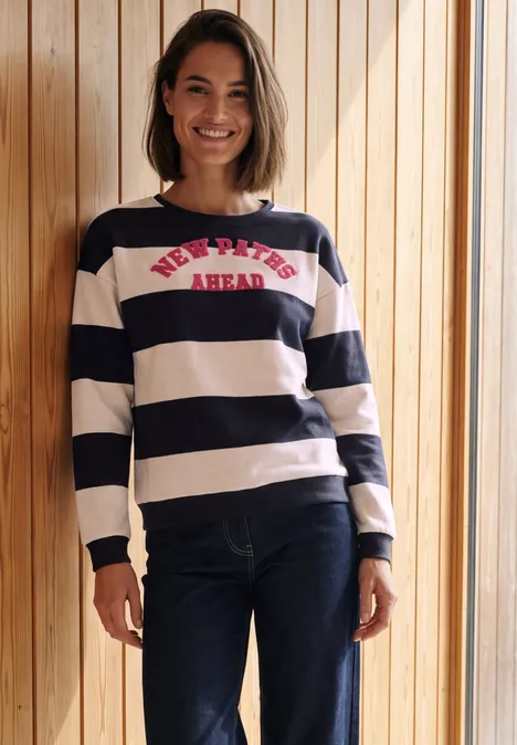 Sweatshirt met strepen en tekstprint urban dark blue Vrouw met marineblauw-wit gestreepte trui en spijkerbroek lacht voor een houten wand.