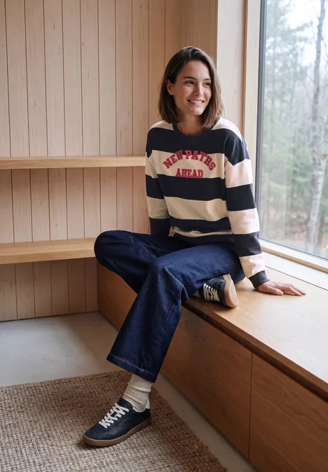 Sweatshirt met strepen en tekstprint urban dark blue Een vrouw lacht terwijl ze ontspannen in een gestreepte trui op een houten vensterbank zit.