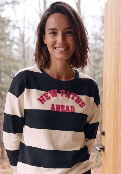 Sweatshirt met strepen en tekstprint urban dark blue Glimlachende vrouw in zwart-wit gestreept shirt met rode tekst NEW PATHS AHEAD voor een houten pilaar buiten.