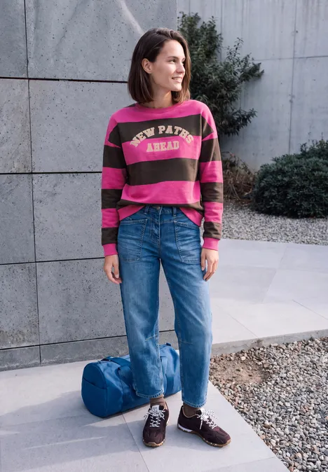 Een vrouw staat voor een betonnen wand, in een gestreepte trui, wijde jeans, loafers en tas naast haar.