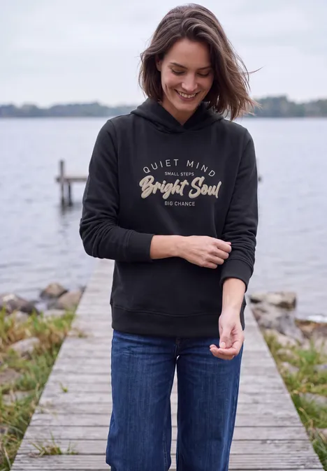 Eine lächelnde Frau in Jeans und schwarzem Kapuzenpullover steht auf einem Steg am Wasser.