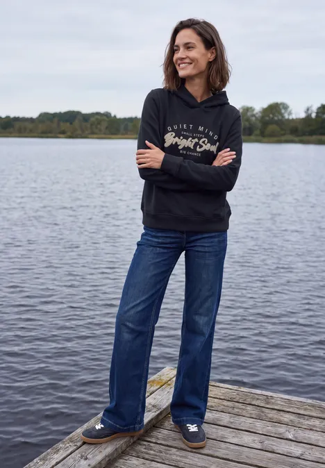 Eine Frau in schwarzer Kapuzenjacke und blauen Jeans steht lächelnd auf einem Holzsteg am Wasser.
