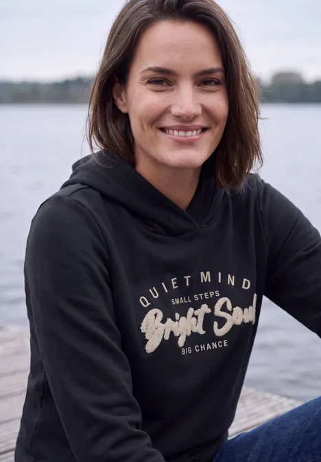 Eine lächelnde Frau in schwarzem Hoodie sitzt entspannt an einem Seeufer.