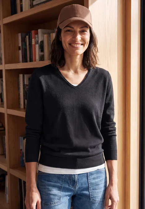 Eine lächelnde Frau in Jeans und Pullover mit Cap vor einem Bücherregal.