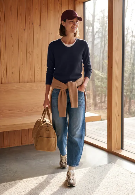 Frau steht in modernem Holzraum am Fenster, trägt Jeans, Pullover, Sneakers und Cap, hält Tasche.
