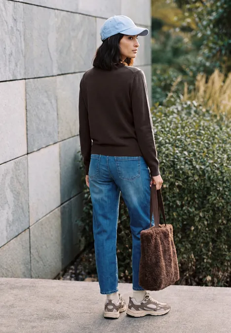 Frau in Jeans, dunkelbraunem Pullover und Cap mit flauschiger Handtasche, steht vor einer Mauer und grünen Büschen.
