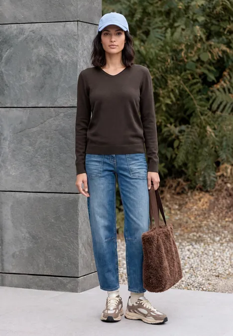Frau mit blauer Mütze, braunem Pullover, blauen Jeans, Sneakern und flauschiger Tasche vor Mauer und Pflanzen.
