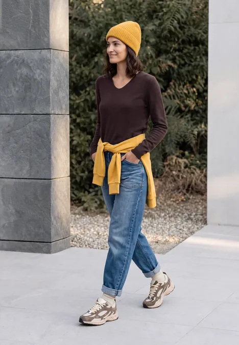 Femme en bonnet jaune, pull marron, jean et baskets marche en souriant en plein air.