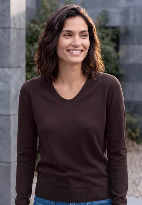 Une femme souriante aux cheveux foncés, portant un pull marron, posant devant un mur de pierre en plein air.