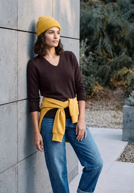 Femme appuyée contre un mur de béton, pull marron, jean, bonnet jaune et sweat noué à la taille.