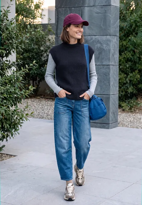Frau in Jeans, Weste und Cap läuft mit blauer Tasche und Sneakers draußen spazieren.