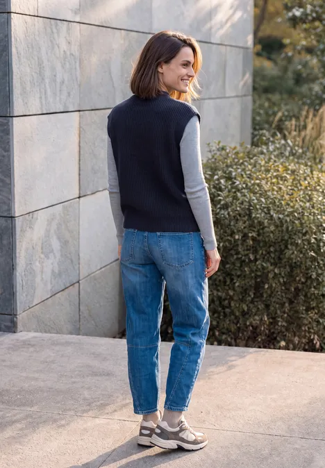Eine Frau steht seitlich vor einer Steinmauer und trägt Jeans, Sneaker und einen ärmellosen Pullover.