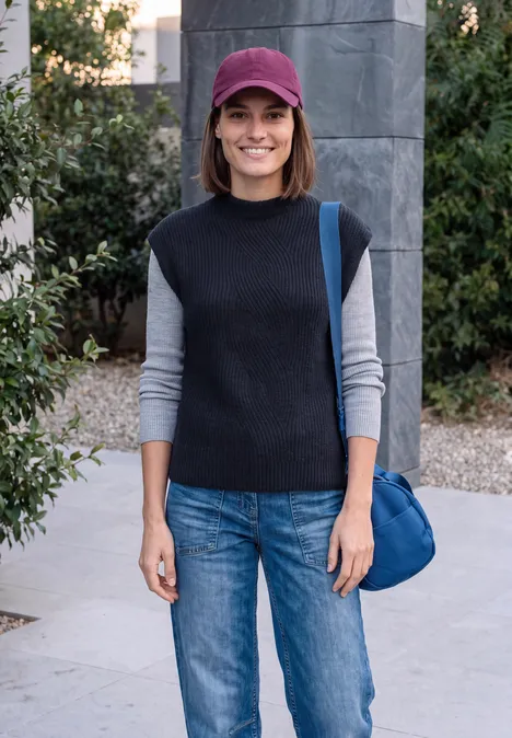 Frau mit roter Mütze, schwarzem Oberteil und Jeans lächelt im Freien vor Gebäudefassade.