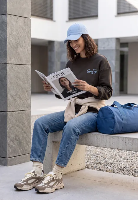 Een vrouw met pet leest rustig een tijdschrift bij een modern gebouw, naast haar blauwe reistas.