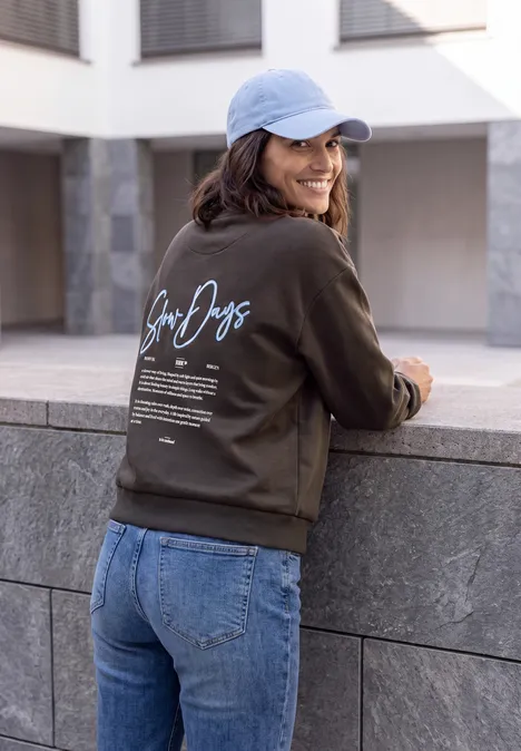 Een vrouw in spijkerbroek en donkergroen sweatshirt met opschrift, lichtblauwe pet, leunt glimlachend tegen een stenen muurtje.