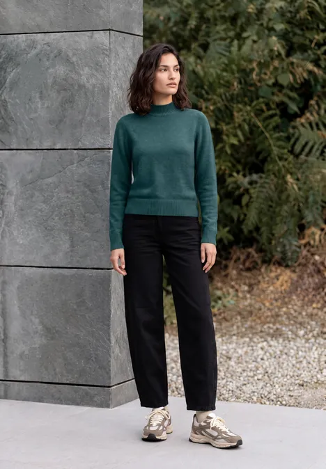 Femme décontractée en pull vert, pantalon noir et baskets, debout devant un mur de béton gris en plein air.