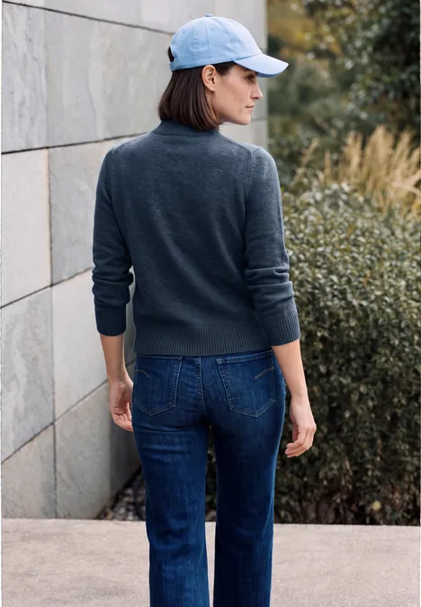 Femme en pull gris, jean et casquette, dos tourné, debout dehors près d'un mur et de buissons.