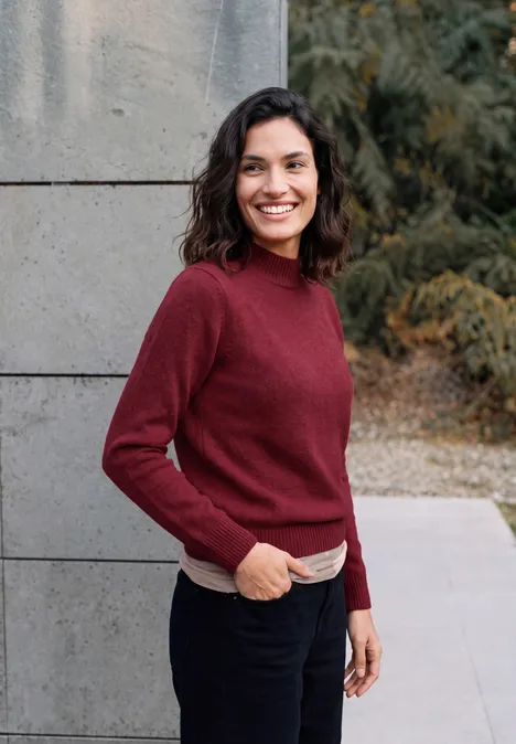 Une femme souriante aux cheveux bruns et pull rouge se tient devant un mur gris en plein air.