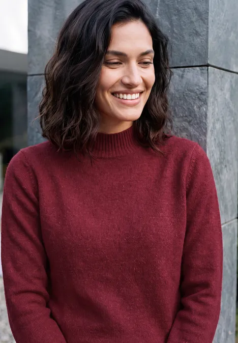 Une femme souriante en pull rouge appuyée contre un mur gris.