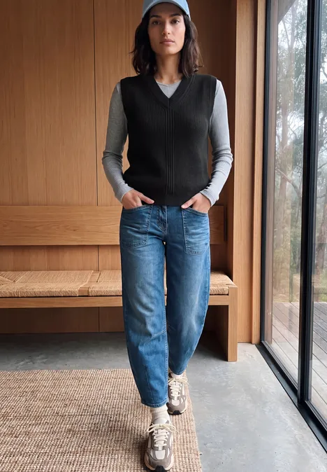 Frau in blauer Mütze, grauem Shirt, schwarzer Weste und Jeans steht vor Holzbank am Fenster.