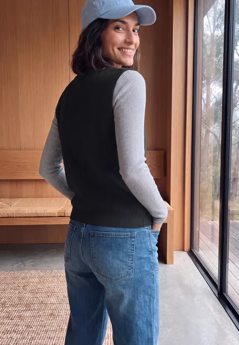 Junge Frau in Jeans, grauem Shirt und schwarzer Weste, mit rückwärts getragener Cap, lächelnd am Fenster.
