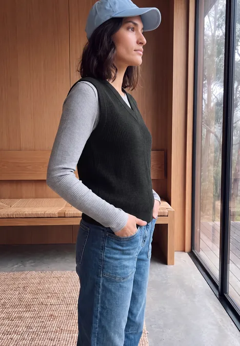 Eine Frau mit Cap, grauem Shirt, schwarzer Weste und Jeans steht seitlich am Fenster.
