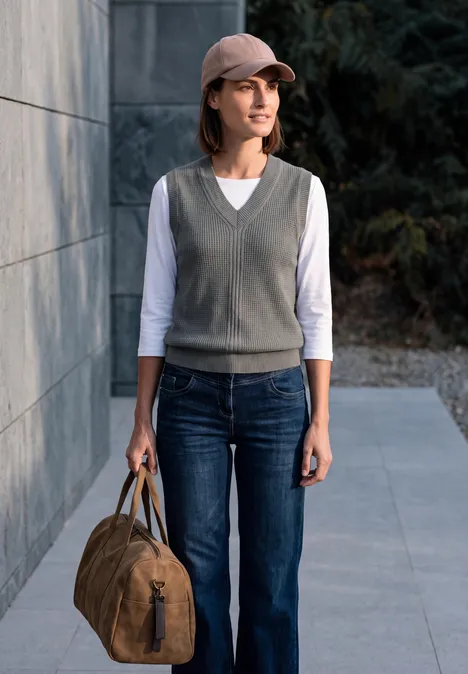 Frau in Jeans, grauer Weste und Cap steht mit brauner Reisetasche vor einer Hauswand.