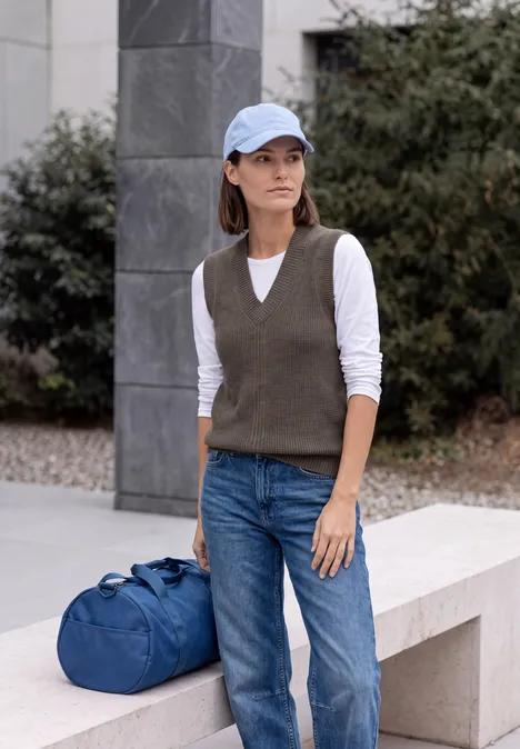 Pullunder in Unifarbe mit V-Neck mushroom taupe melange Frau mit Baseballcap, Strickweste, Jeans und blauem Reisetasche vor urbaner Kulisse.