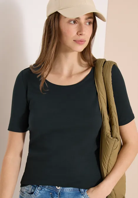 Une femme porte un t-shirt noir à manches courtes, un jean, une casquette et un sac épaule vert matelassé.