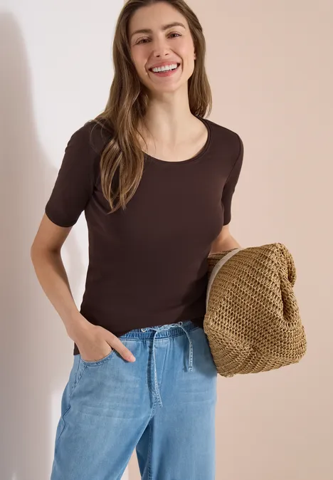 Vrouw in bruin T-shirt en spijkerbroek houdt een lichtbruin, grofgebreide tas vast.