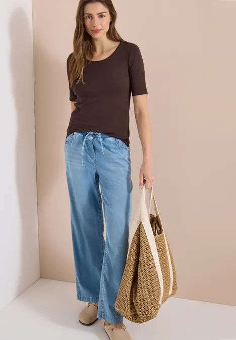 Vrouw in wijde denim, donker T-shirt, met strozak en lichte schoenen voor een beige muur.