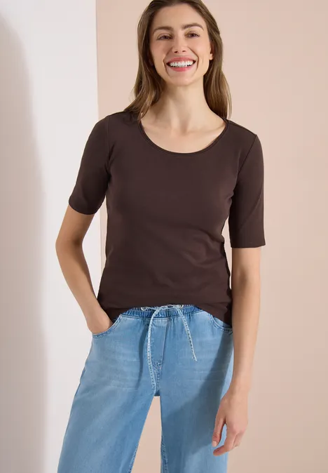 Vrouw draagt een donkerbruin T-shirt en lichtblauwe jeans met trekkoord, staand voor een lichte achtergrond.