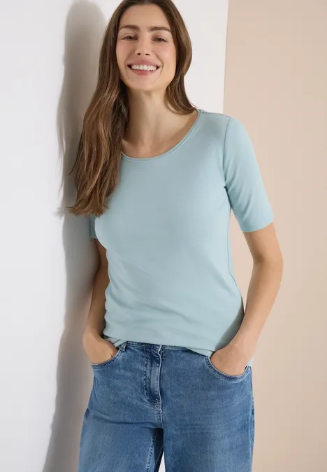 Vrouw leunt tegen een muur, draagt een lichtblauw t-shirt en een spijkerbroek.