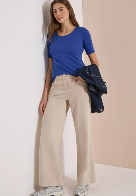 Basic T-Shirt mit Rundhals in Unifarbe deep river blue Frau trägt enges blaues Shirt und weite beigefarbene Hose, hält dunkelblaue Jacke lässig.