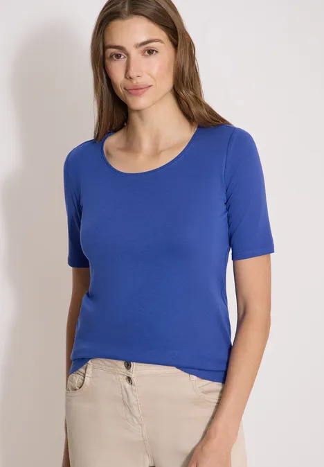 Basic T-Shirt mit Rundhals in Unifarbe deep river blue Junge Frau in blauem Kurzarmshirt und beiger Hose vor weißem Hintergrund.