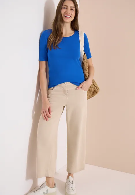 Vrouw leunt tegen muur, draagt blauwe top, beige wijdvallende broek, witte sneakers en rieten tas.