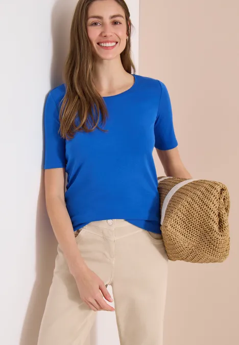 Vrouw leunt tegen lichte muur, draagt blauw T-shirt, beige broek en houdt een strohoed vast.