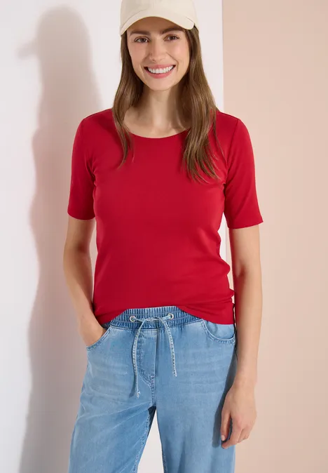 Vrouw voor een lichte muur, met rood shirt en blauwe jeans, handen in haar zakken.