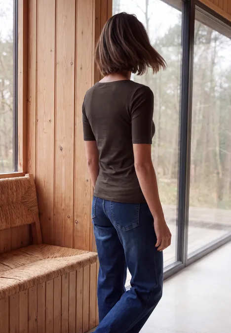 Eine Frau im grünen T-Shirt und Jeans steht mit dem Rücken zur Kamera vor großen Fenstern in einem Holzraum.