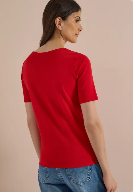 Eine Frau von hinten in rotem T-Shirt und blauer Jeans vor neutralem Hintergrund.